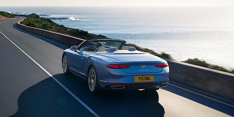 Bentley Barcelona New Bentley Continental GTC Azure convertible rear view in Light Windsor blue paint with 22 inch ten spoke wheels driving dynamically on an ocean road in the sunshine