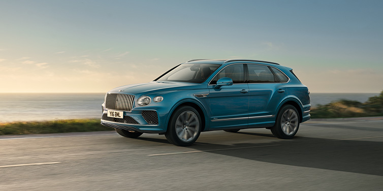 Bentley Barcelona Bentley Bentayga Azure SUV in Topaz blue paint driving dynamically by the ocean with 22 inch 10 spoke directional wheels