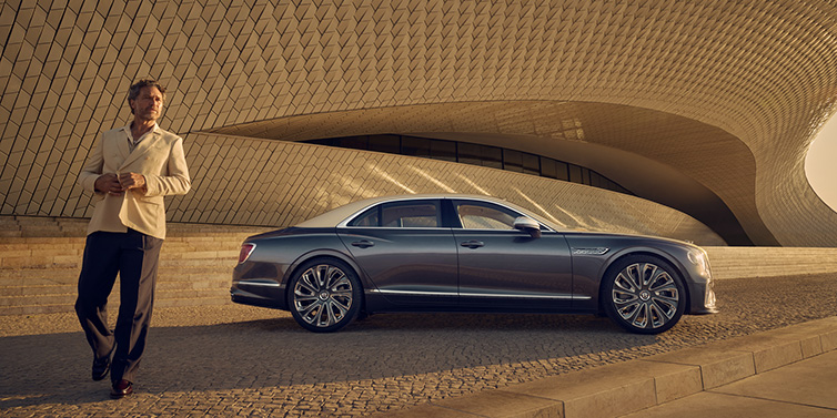 Bentley Barcelona Bentley Flying Spur Mulliner sedan in Rose Gold over Magnetic duotone paint parked by a curved wall with person in suit jacket standing on the left